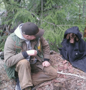 Image of Man in Extreme Cruiser Forestry Vest Sitting Next To Dog In A Raincoat